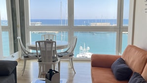 A living room with a large window overlooking a marina. There is a round table with chairs, a sofa, and a coffee table with a remote control.