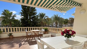 Eine Terrasse mit weißem Tisch und Stühlen, Topfpflanzen und Blick auf Palmen und Gebäude.