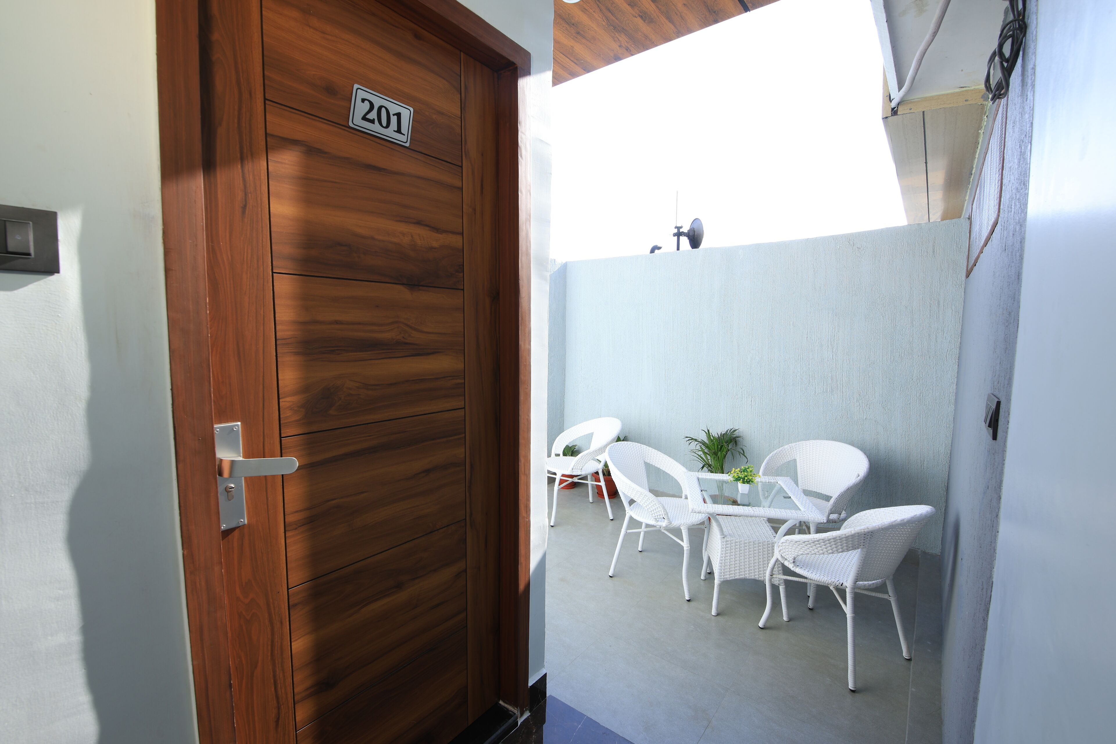 Puerta de madera con placa de número, sillas de mimbre blanco y una mesita en un balcón.