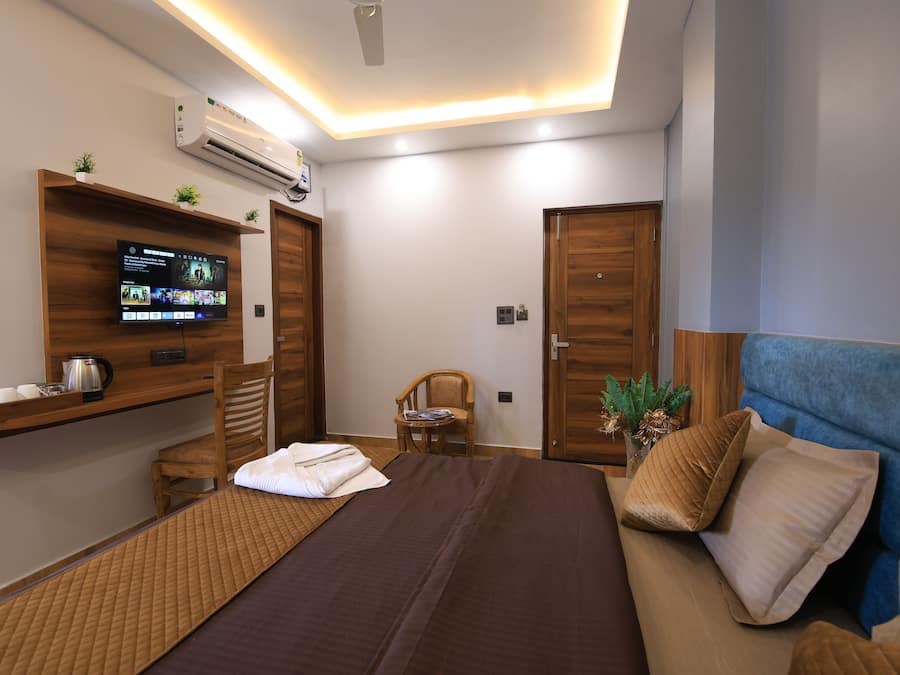 Una habitación de hotel moderna con una cama, un televisor montado en la pared, un estante de madera con artículos de baño, una silla y un sofá azul.