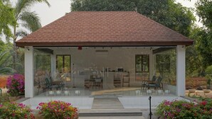 A covered outdoor seating area with glass walls, surrounded by lush greenery and flowering plants. - Rocks and Greens (Padmanabham)