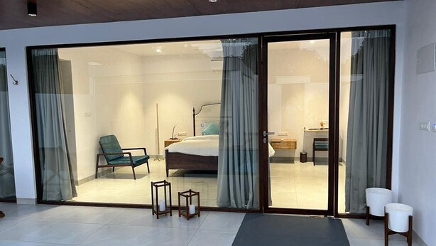 A modern hotel room with a large sliding glass door leading to a balcony, a bed, a chair, and a small table with candles. - Rocks and Greens (Padmanabham)