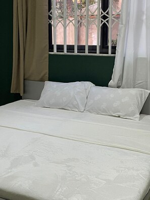 A bed with white linens and a green headboard, a window with white bars, and beige curtains. - Serene Hostel  Accra (Accra)