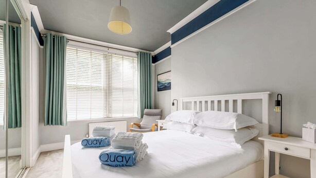 Ein ordentlich bezogenes Bett mit weißen Leinen und einem blauen Handtuch mit dem Aufdruck "Quay", ein Nachttisch mit Lampe und ein Fenster mit grünen Vorhängen.