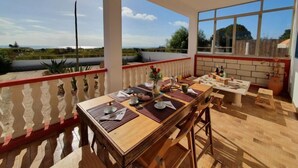 Un coin repas avec une longue table en bois dressée pour deux personnes, une grande fenêtre donnant sur la campagne et un balcon agrémenté d’une balustrade.