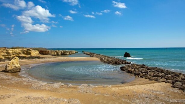 Une plage avec une eau d’un bleu limpide, un rivage rocheux et une zone sablonneuse où se trouve un petit bassin d’eau.