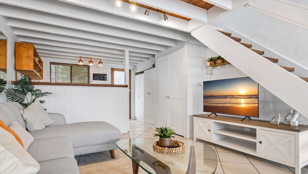 A modern living room with a glass coffee table, a sofa, a TV on a wooden stand, and a staircase with a sunset view.