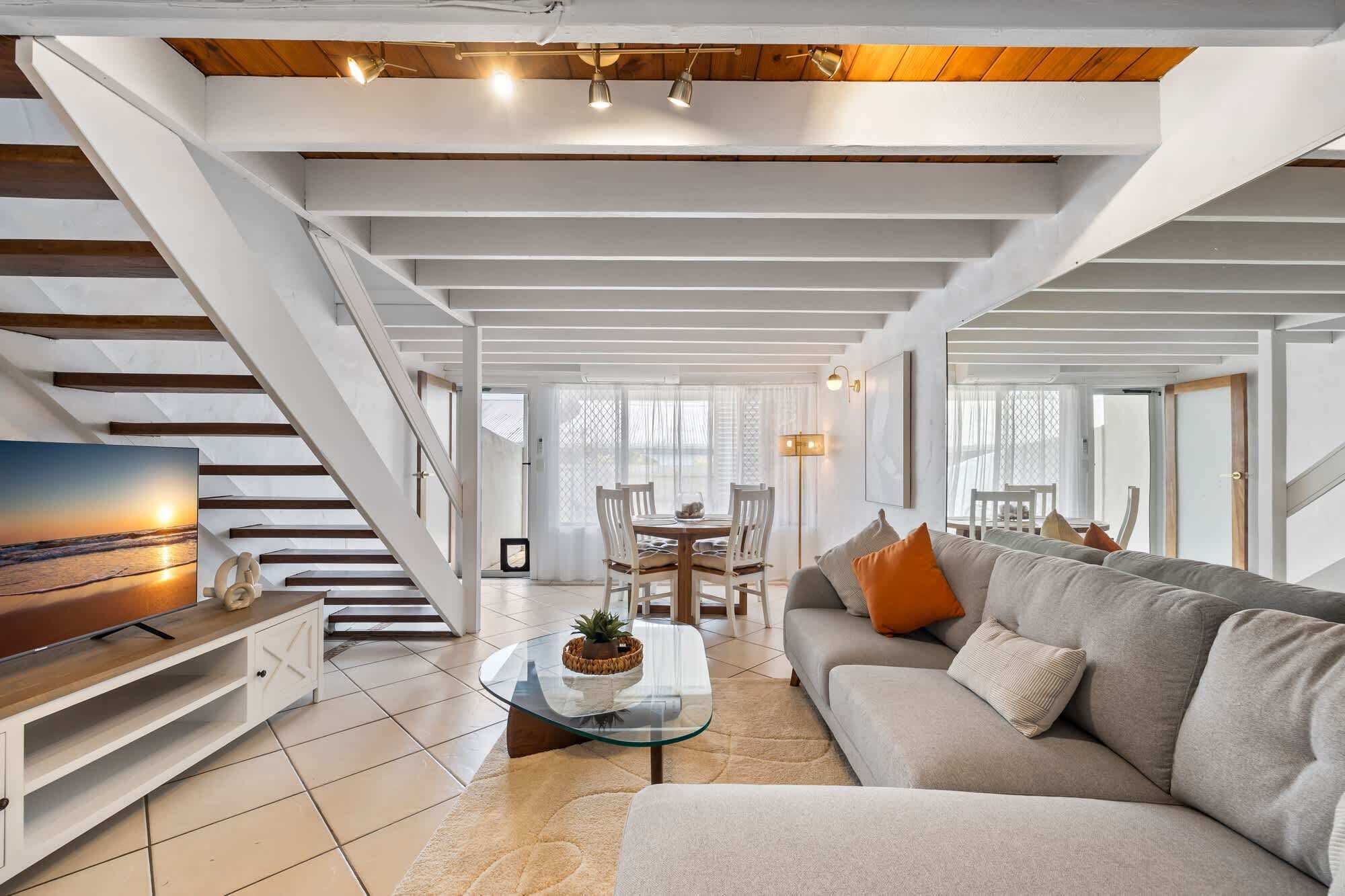 A spacious living room with a staircase, a glass coffee table, a sofa, and a TV on a wooden cabinet.