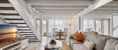 A spacious living room with a staircase, a glass coffee table, a sofa, and a TV on a wooden cabinet.