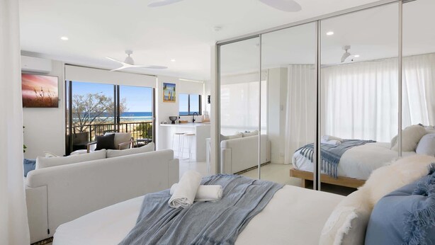Ein modernes Hotelzimmer mit einem großen Bett, einem Balkon mit Meerblick, einem Essbereich und einem großen Spiegel, der den Raum reflektiert.