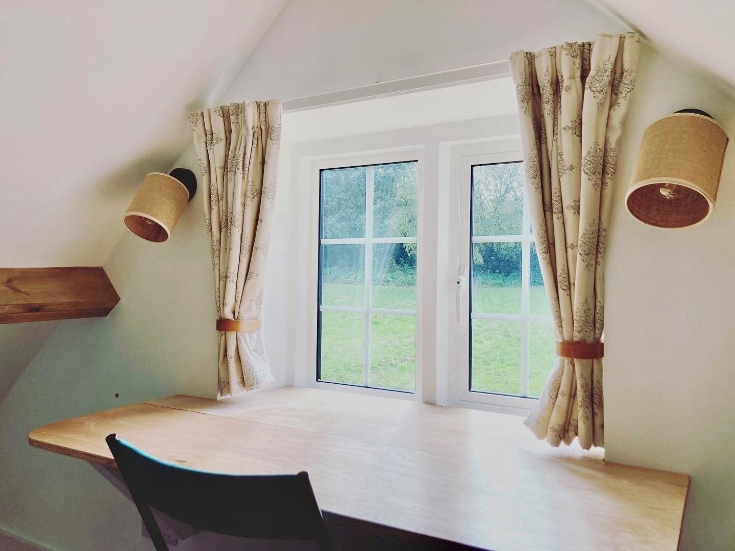A room with a wooden table and a chair, a window with curtains, and a lamp on the wall.