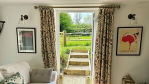 A cozy living room with a sofa, a coffee table with newspapers, and a view of a garden through a glass door.