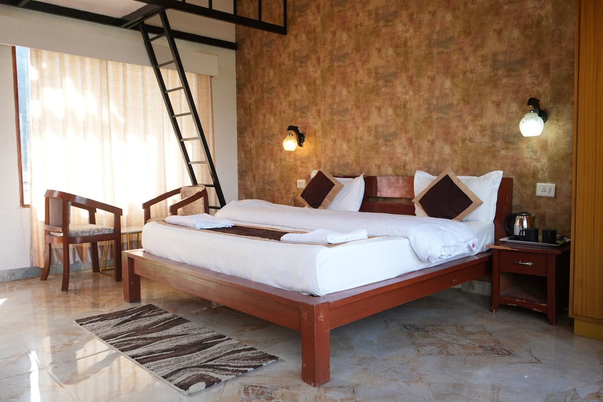 A spacious bedroom with a large bed, wooden headboard, and a bench. There is a chair, a nightstand, and a lamp on the wall.