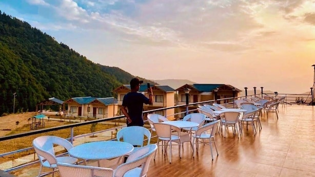 A terrace with white chairs and tables, overlooking a forested hillside and a row of cabins. - Resort Prakriti Unwind (Chakrata)