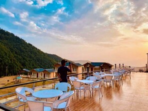 A terrace with white chairs and tables, overlooking a forested hillside and a row of cabins. - Resort Prakriti Unwind (Chakrata)