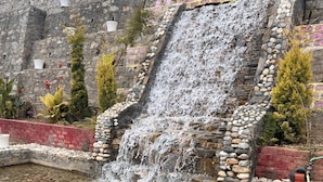 A stone waterfall cascading into a pebble-lined pool, surrounded by a stone wall with plants and a brick base. - Resort Prakriti Unwind (Chakrata)