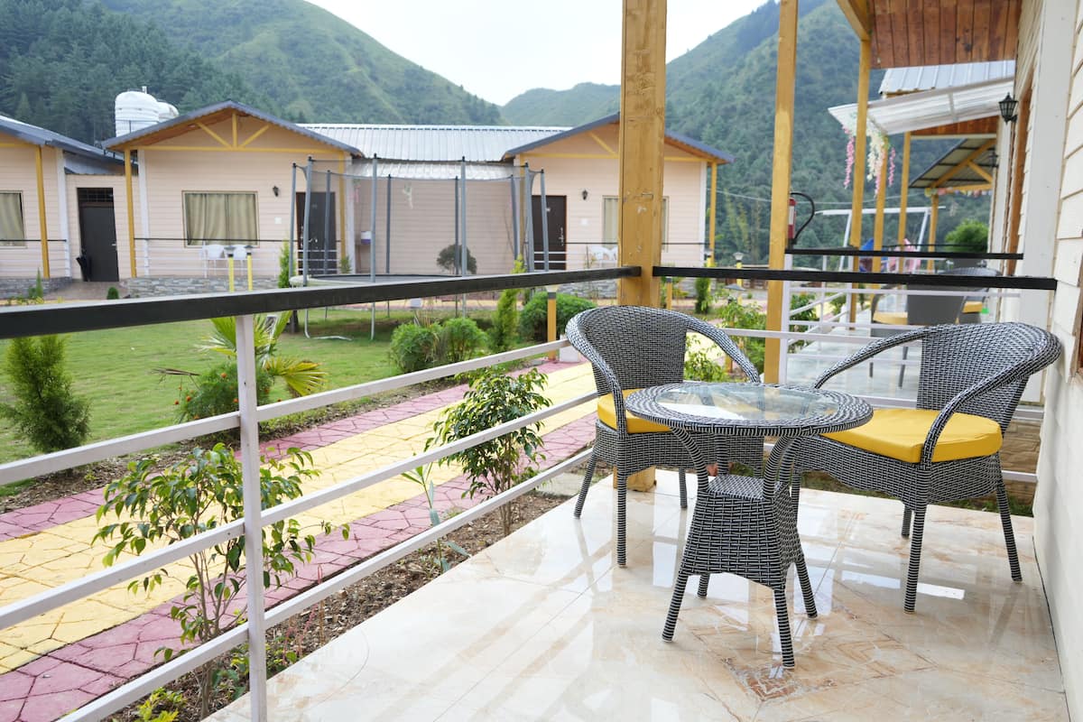 A balcony with wicker chairs and a glass table, overlooking a garden and buildings.