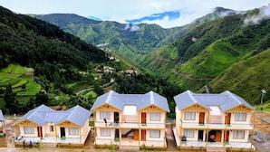A row of small, single-story cabins with sloped roofs, set against a backdrop of lush green hills and a valley. - Resort Prakriti Unwind (Chakrata)
