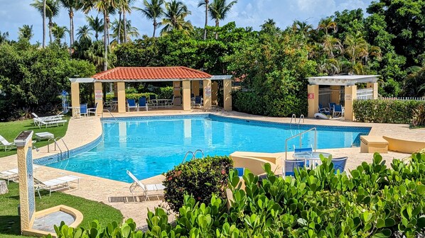 A swimming pool with lounge chairs, a covered seating area, and lush greenery in the background. - 2Bed/2Bath at the Tennis Village at Palmas del Mar (Humacao)