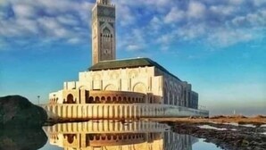 Un edificio grande y rectangular con una torre central, rodeado por una masa de agua tranquila que refleja su estructura.