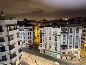 A multi-story building with balconies, a street in front, and a cafe with a sign in a foreign language.