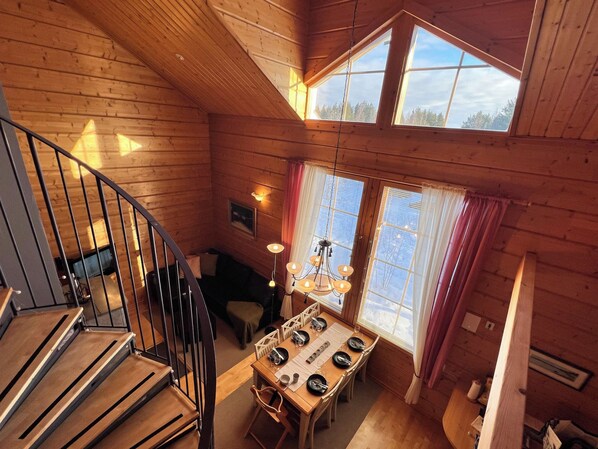 A wooden cabin interior with a dining area, a staircase, and a large window.