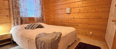 A bedroom with a bed, a bedside table, a window with curtains, and a wooden wall.