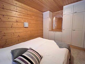 A bedroom with a wooden ceiling, a bed with a striped pillow, a white wardrobe, and a mirror above the wardrobe.