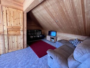 A cozy attic room with a wooden ceiling, a sofa, a red rug, and a television.