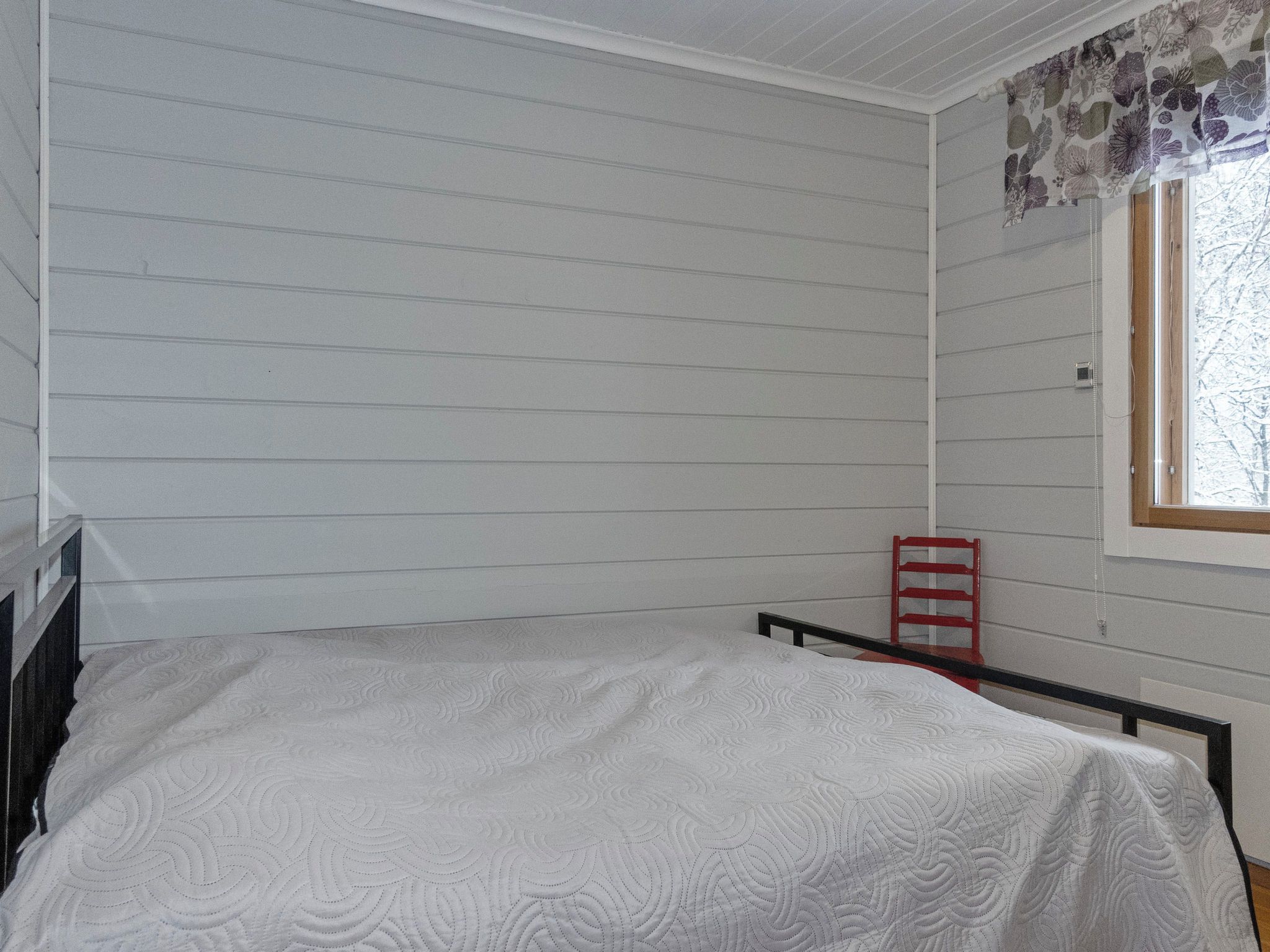 Ein Zimmer mit einem Einzelbett, einem Fenster mit Blumenvorhang und einem roten Sessel.