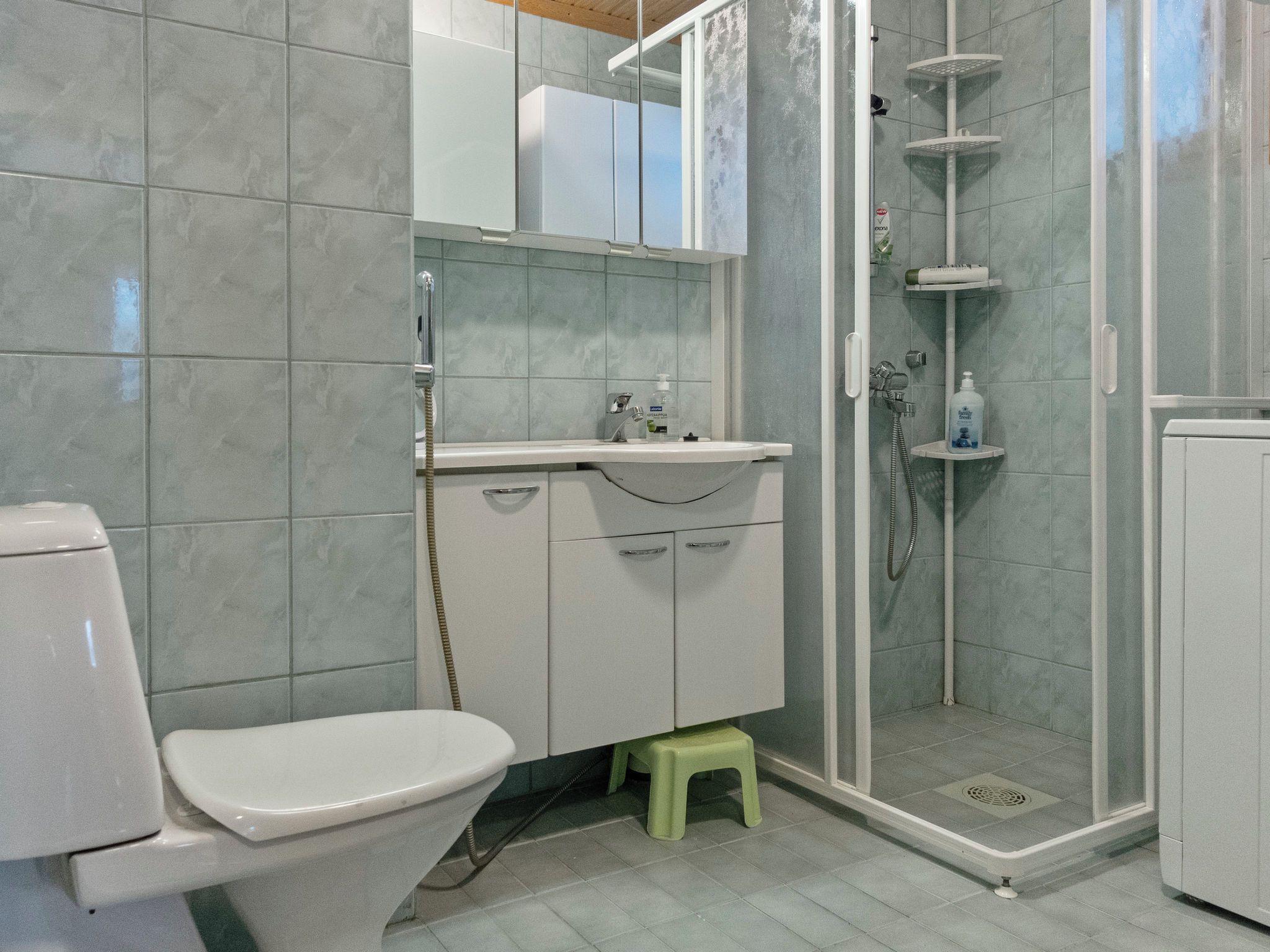 A bathroom with a toilet, a sink with a mirror, a shower enclosure, and a stool.