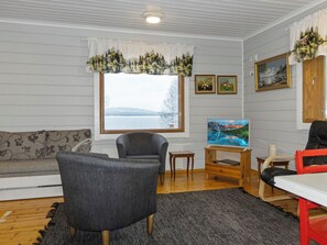 A living room with a sofa, armchairs, a TV on a wooden stand, a window with a view of a lake, and a door with a painting.