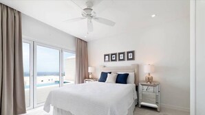 A bedroom with a large bed, a ceiling fan, a nightstand with a lamp, and a view of the ocean through a sliding door. - White 4 bedroom villa with amazing ocean views in a gated community in St James (Fitts Village)