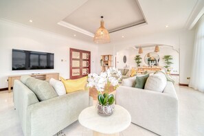 A modern living room with a sofa set, a coffee table with orchids, a flat-screen TV, and a dining area in the background.