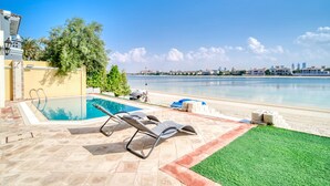 Une piscine avec des chaises longues, une pelouse et une vue sur la mer, avec des bâtiments visibles au loin.