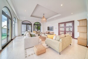 A spacious living room with large windows, a modern sofa set, a wooden bookshelf, and a flat-screen TV.