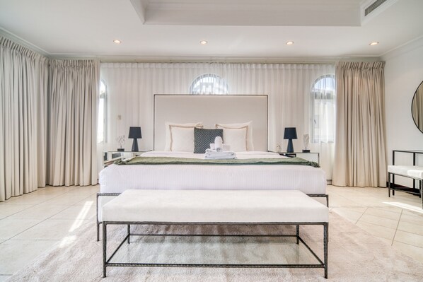 A large, well-lit bedroom with a large bed, two bedside tables, a bench, and a round mirror on the wall.