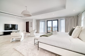 A modern bedroom with a large bed, a flat-screen TV, a white armchair, and a view of the city through large windows.