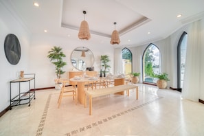 A spacious dining area with a wooden table set for six, surrounded by chairs. There are hanging woven light fixtures, a round mirror on the wall, and potted plants.