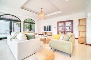 A spacious living room with two sofas, a coffee table, and a TV. There is a large window with a view of greenery outside.