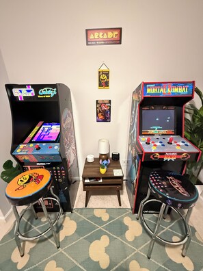 Dos máquinas de videojuegos, una lámpara, una mesita y una planta en una habitación con alfombra estampada.