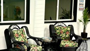 Deux chaises en osier avec un revêtement à motifs floraux, une petite table et une plante en pot, devant un bâtiment doté de grandes fenêtres et d’un mur blanc.
