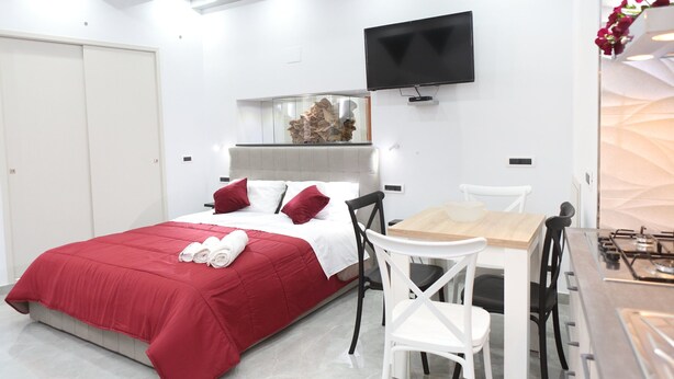 Ein modernes Hotelzimmer mit Bett, Wandfernseher, einem kleinen Esstisch mit Stühlen und einer Küchenzeile mit Herd.