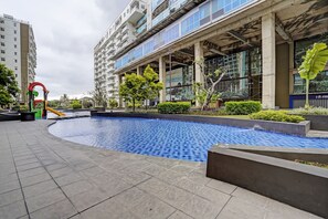 Une piscine dotée d’une terrasse carrelée, entourée de verdure et bordée d’un bâtiment moderne avec de grandes fenêtres et des balcons.