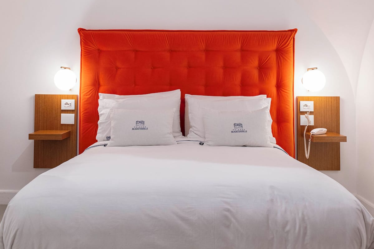 Una habitación de hotel con una cama grande, dos almohadas blancas, un cabecero tapizado en rojo y lámparas de noche.