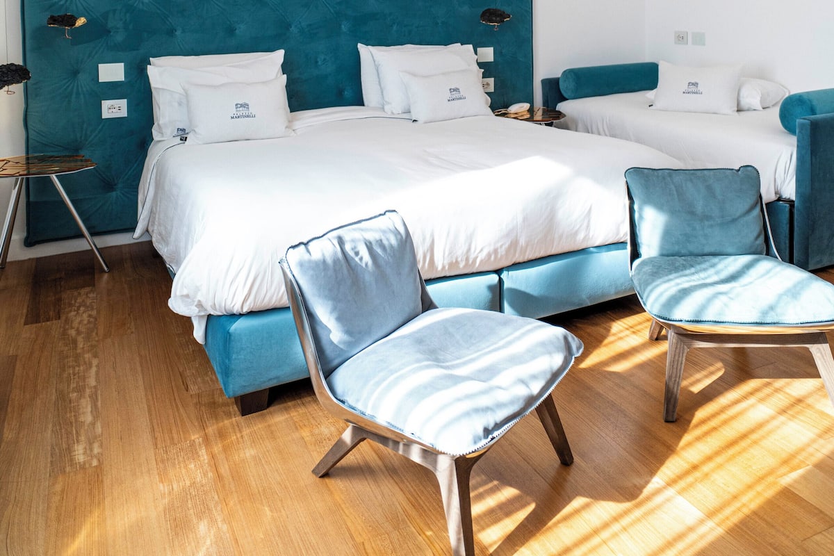 Una habitación de hotel moderna con una cama grande, dos camas más pequeñas, un rincón de descanso con sillones y un espejo instalado en el techo.