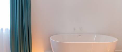 A modern bathroom with a large white bathtub, wooden flooring, and teal curtains.