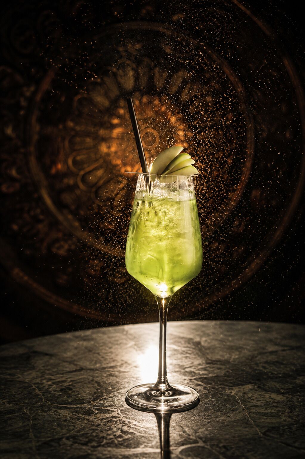 A cocktail glass with a green drink, garnished with an apple slice, on a marble surface.