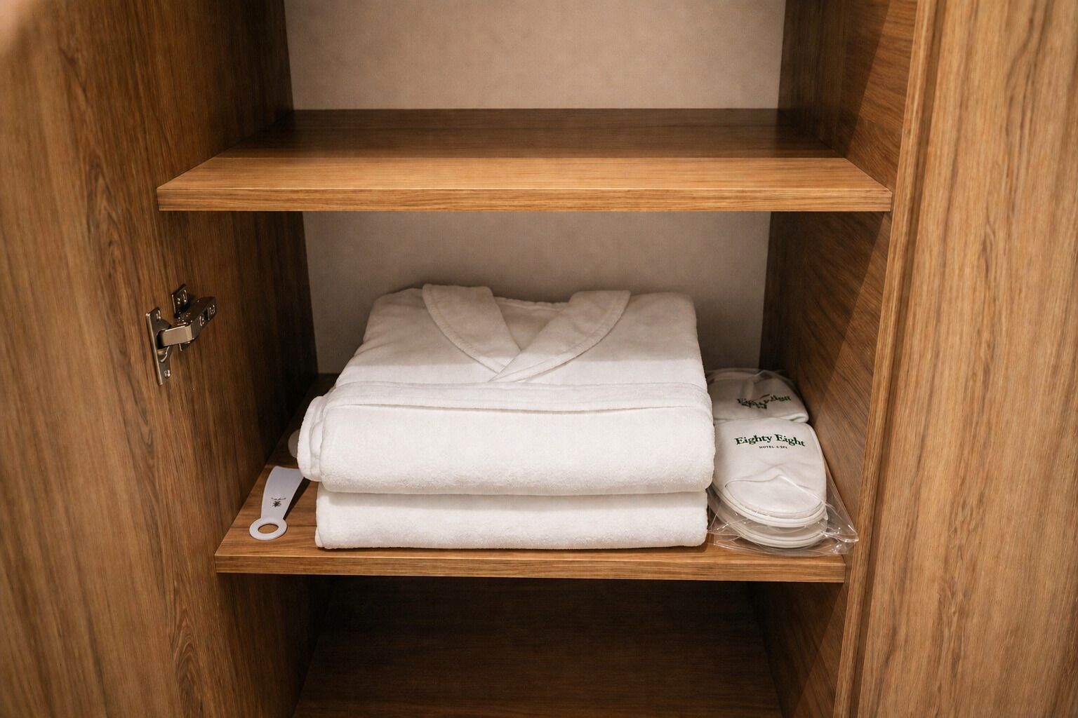 A wooden wardrobe with two shelves; the top shelf holds a folded white robe, and the bottom shelf has a folded white towel and a plastic bag with a tag.