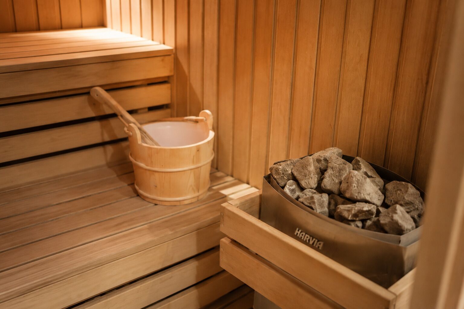A sauna with wooden benches, a wooden bucket, and a metal container filled with rocks.
